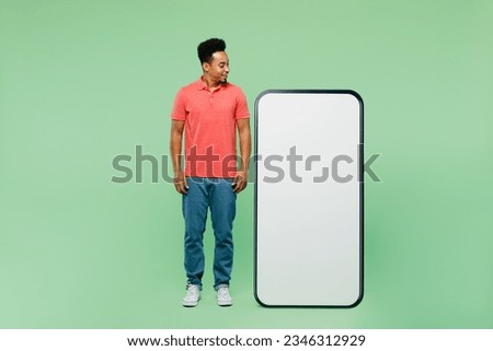 Full body smiling happy fun young man wear pink t-shirt look at big huge blank screen mobile cell phone smartphone with area isolated on plain light green background studio portrait. Lifestyle concept