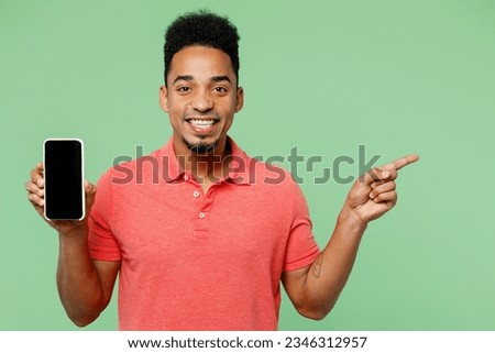Young man of African American ethnicity wears pink t-shirt hold in hand use mobile cell phone with blank screen workspace area point finger aside isolated on plain pastel light green background studio