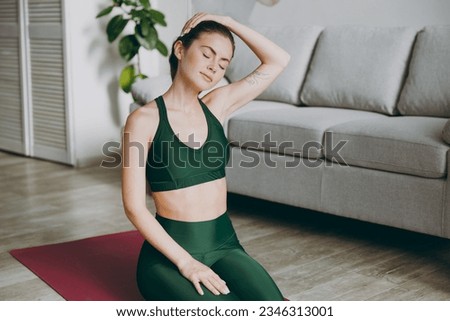 Young caucasian strong sporty athletic fitness trainer instructor woman wear green tracksuit holding head move it close eyes training do exercises at home gym indoor. Workout sport motivation concept Royalty-Free Stock Photo #2346313001