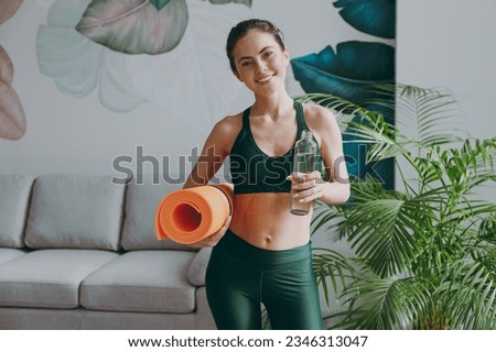 Young fun strong sporty athletic fitness trainer instructor woman wearing green tracksuit hold in hand yoga mat drink water training do exercises at home gym indoor. Workout sport motivation concept Royalty-Free Stock Photo #2346313047