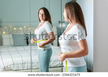Smiling young woman after weight loss measuring waist in front of mirror Royalty-Free Stock Photo #2346321675
