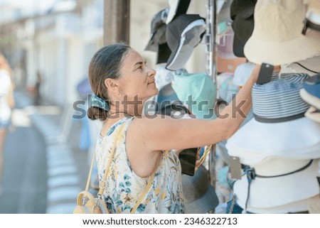 Mature woman choosing and trying on straw hat in street shop in resort town. Summer fashion and shopping idea