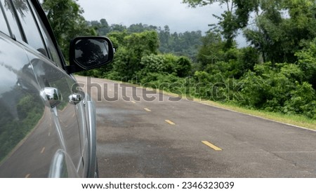 Mirror wing and blurred of beside of grey car. Wet of asphalt road after rain. Beside with green grass and forest under the sky and dark clouds. Travel trip of reservoir of Khiri Than Chanthaburi.