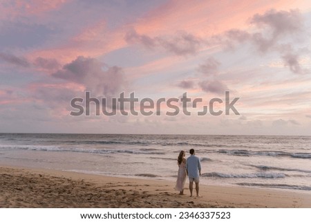 Two people stand on the beach with their backs to the camera, facing the sea during sunset. They gaze into the distance at the pink-hued sunset. The clouds in the sky are also tinged with pink. It's t Royalty-Free Stock Photo #2346337523