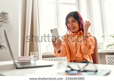 Asian young girl with bright pink strands of hair sitting at a table with a laptop at home in the living room, shocked looking at the smartphone and showing yes gesture. Royalty-Free Stock Photo #2346403085