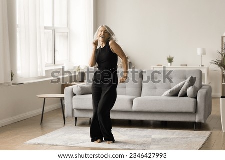 Joyful active senior lady excited with single dance at home enjoying motion, activity, music, singing song, having fun in cozy comfortable living room interior. Full length wide shot