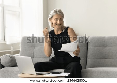 Happy successful mature investor woman celebrating profit, income from investment, retirement savings, reading financial paper document, making winner hand, smiling, sitting at laptop at home Royalty-Free Stock Photo #2346428001