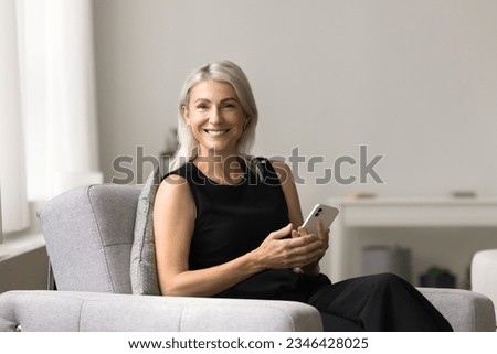 Cheerful attractive mature woman holding white smartphone, resting in soft comfortable armchair, looking at camera, smiling for portrait, promoting useful online application, Internet service