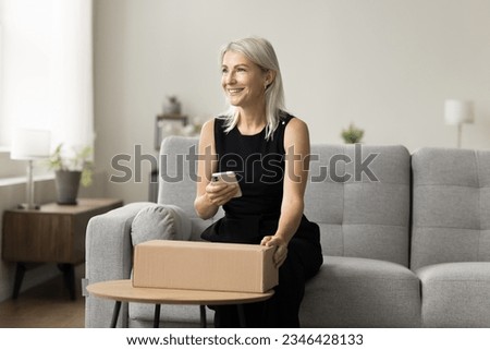 Happy mature delivery service customer woman receiving parcel, using e-commerce logistic app on smartphone, sitting on sofa, looking away with dreamy thoughtful face, thinking