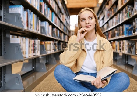 Smart pretty creative girl student holding book sitting on floor among bookshelves in modern university campus library looking away thinking of college course study thinking reading literature.