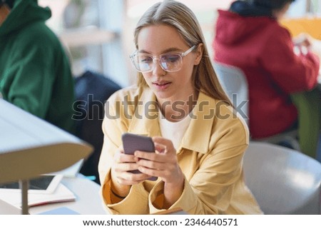 Girl student using mobile phone looking at smartphone sitting at desk in university college campus classroom. Young blonde woman holding cellphone modern tech in university advertising apps. Royalty-Free Stock Photo #2346440571