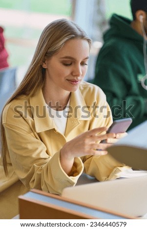 Teen girl gen z student using mobile phone looking at smartphone sitting at desk in university college campus classroom. Young blonde woman holding cellphone modern tech in university, vertical. Royalty-Free Stock Photo #2346440579