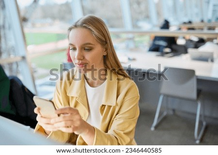Teen girl gen z student using mobile phone looking at smartphone sitting at desk in university college campus classroom. Young blonde woman holding cellphone technoogy in university. Royalty-Free Stock Photo #2346440585