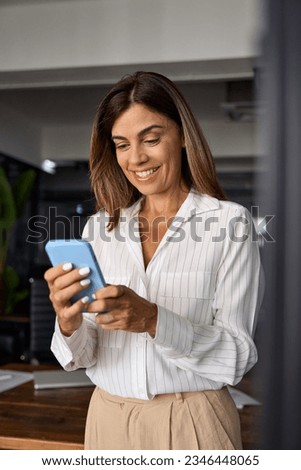Vertical portrait of European businesswoman CEO holding smartphone using fintech application sitting on sofa indoors. Smiling Latin Hispanic mature adult professional business woman using mobile phone Royalty-Free Stock Photo #2346448065