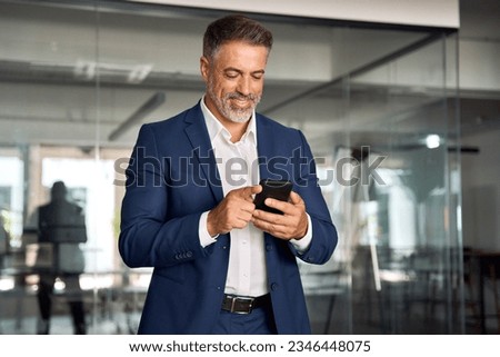 Smiling mature Latin or Indian businessman holding smartphone in office. Middle aged manager using cell phone mobile app. Digital technology application and solutions for business success development. Royalty-Free Stock Photo #2346448075