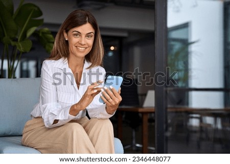 European businesswoman CEO holding smartphone using fintech application sitting on sofa in modern office. Smiling Latin Hispanic mature adult professional business woman using mobile phone cellphone.  Royalty-Free Stock Photo #2346448077