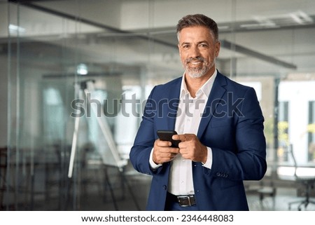 Smiling mature Latin or Indian businessman holding smartphone in office. Middle aged manager using cell phone mobile app. Digital technology application and solutions for business success development. Royalty-Free Stock Photo #2346448083