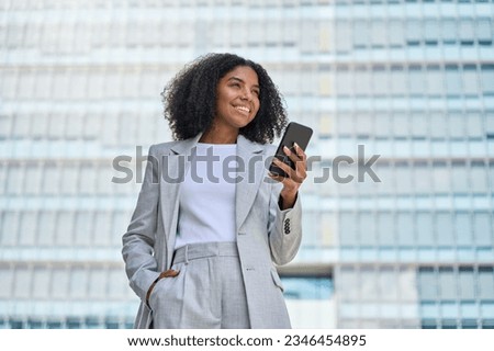 Happy confident young African American business woman using cell mobile phone outdoor. Smiling lady professional holding cellphone standing at city street building looking away working outside office. Royalty-Free Stock Photo #2346454895