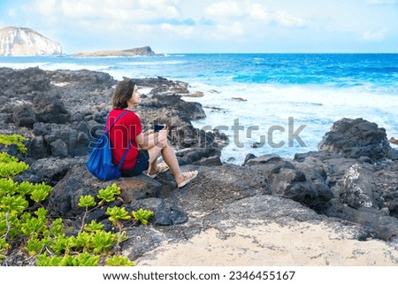 Young woman sitting on lavarocks along the Hawaiian ocean at Makapu'u beach taking pictures with cellphone