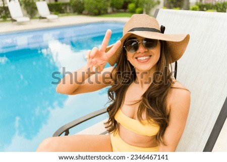 Latin happy young woman making the peace sign and smiling while looking at the camera relaxing by the pool in the summer