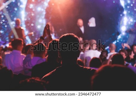 A crowded concert hall arena with scene stage lights with musicians band on a stage at the venue, rock show performance, with concert-goers attendees, audience on dance floor during concert festival
