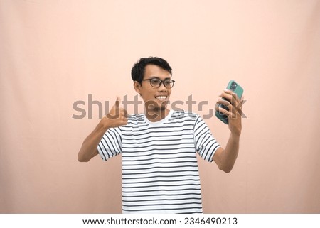 portrait of an asian man wearing glasses wearing a casual striped t-shirt. Posing showing thumbs up, recommending, okay, agreeing and holding smartphone. Isolated beige background.