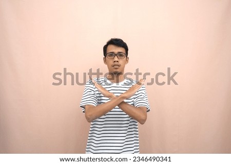 portrait of an asian man in glasses wearing a casual striped t-shirt. denote stop, cross prohibition sign, prohibit something, refuse,. beige background.