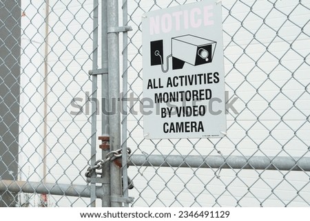 all activities monitored by video camera white rectangle sign with black print and illustration picture of video camera against silver diamond fence
