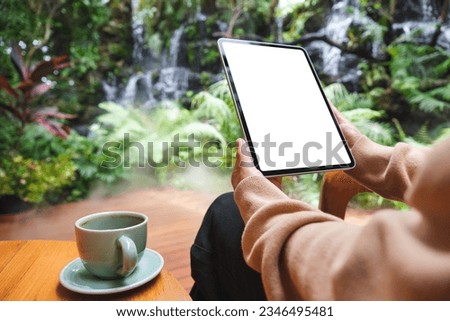 Mockup image of a woman holding digital tablet with blank white desktop screen in the garden with waterfall