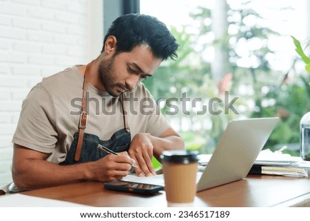 Indian man being barista and coffee shop owner sitting at desk Take some serious thought into checking accounts. manage cost of small coffee shop. Check accounting documents with laptop computer