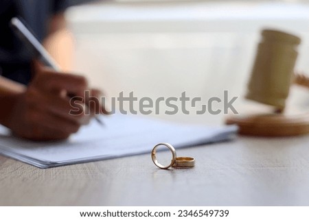 Wedding rings on wooden table with hand signing documents and judge gavel on the background. Marriage and divorce concept Royalty-Free Stock Photo #2346549739