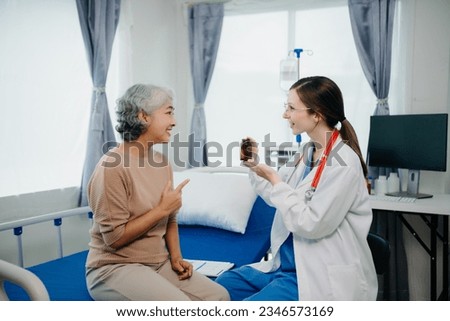 Caucasian doctor in white suit take notes while discussing and Asian elderly, woman patient who lying on bed with receiving saline solution in hospital or clinic.
