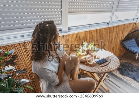 Young caucasian woman in a white shirt sitting on a balcony with roller shutters. Sun protection and heat  Royalty-Free Stock Photo #2346609969