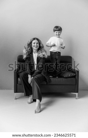 Mom and son at a photo shoot in the studio. Mom is sitting on the sofa. The boy is standing and smiling. Mom is wearing a black pantsuit. The boy is wearing a white T-shirt. Black  white photo.