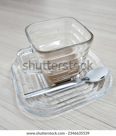 a photography of a glass cup of coffee on a plate with a spoon, there is a glass cup of coffee on a plate with a spoon.