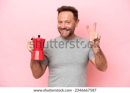 Middle age caucasian man holding coffee pot isolated on pink background smiling and showing victory sign