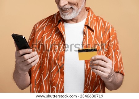 Closeup of smiling gray haired senior man wearing stylish shirt holding mobile phone and credit card isolated on beige background. Elderly man paying. Concept of mobile banking