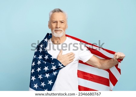 Portrait of attractive senior american man holding hand on heart, american flag, patriot wearing white t shirt isolated on blue background. Handsome retired man posing for picture in studio