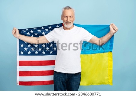 Portrait of smiling positive senior man wearing white t shirt holding American and Ukrainian flags isolated on blue background, mockup, copy space. Concept of support, patriotism, union