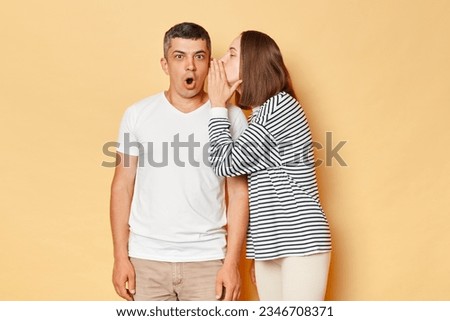 Shocked man hearing gossips from woman talkign to his ear posing with widely opened mouth being amazed and susrprised wearing casual clothing standing isolated over beige background Royalty-Free Stock Photo #2346708371