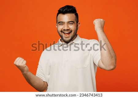 Young happy overjoyed Indian man he wears white t-shirt casual clothes doing winner gesture celebrate clenching fists say yes isolated on plain orange red background studio portrait. Lifestyle concept Royalty-Free Stock Photo #2346743413