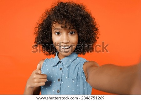 Close up of amazed little african american kid girl 12-13 in denim dress doing selfie shot on mobile phone point on camera isolated on orange background studio portrait. Childhood lifestyle concept