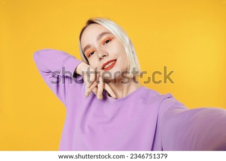 Smiling young blonde caucasian woman 20s bob haircut bright makeup wearing casual basic purple shirt close up doing selfie shot on mobile phone isolated on yellow color background studio portrait