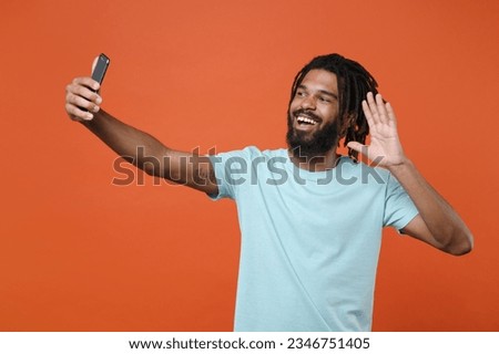 Cheerful young african american man guy wearing blue casual t-shirt posing isolated on orange background studio. People lifestyle concept. Doing selfie shot on mobile phone waving greeting with hand