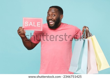 Funny man of African American ethnicity 20s he wears casual pink t-shirt Hold package bag with purchases after shopping, sign with SALE title isolated on pastel plain light blue color wall background