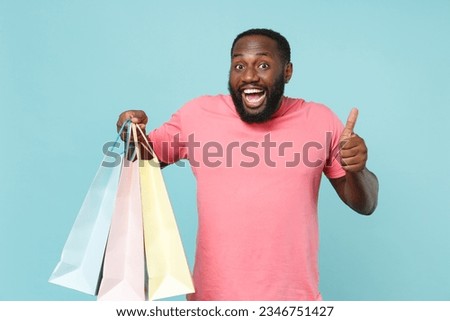 Excited man of African American ethnicity 20s he wears casual pink t-shirt holding package bags with purchases after shopping isolated on pastel plain light blue color wall background studio portrait