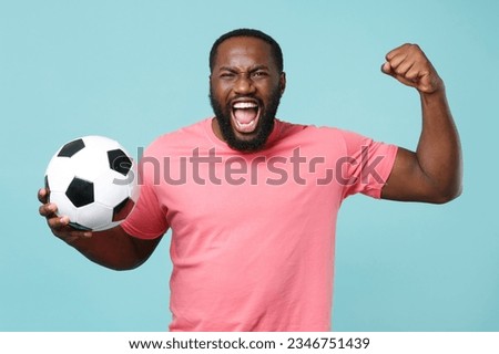 Fun man of African American ethnicity 20s fan he wear pink t-shirt cheer up support football sport team hold in hand ball watch tv live stream isolated on pastel plain light blue color wall background