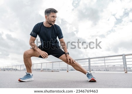 A trainer in fitness clothes is a T-shirt, running sports shoes and a smart watch for sports.    A strong man. A confident male athlete training does warm-up exercises.
