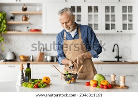 Happy senior man preparing healthy vegetable salad in kitchen, seasoning meal, smiling elderly gentleman in apron adding salt to bowl, enjoying cooking vegetarian food at home, free space