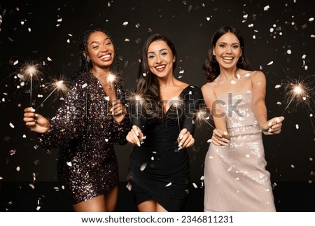 Pretty multiethnic young women wearing nice elegant outfits holding bengal lights, laughing among confetti sparkles, friends celebrating New Year 2024 together, isolated on black background Royalty-Free Stock Photo #2346811231